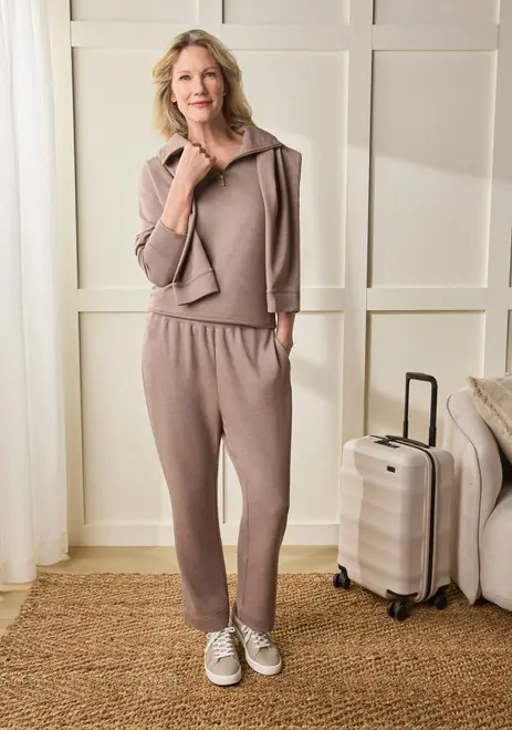 Woman over 50 wearing a taupe knit travel set with sneakers and rolling luggage