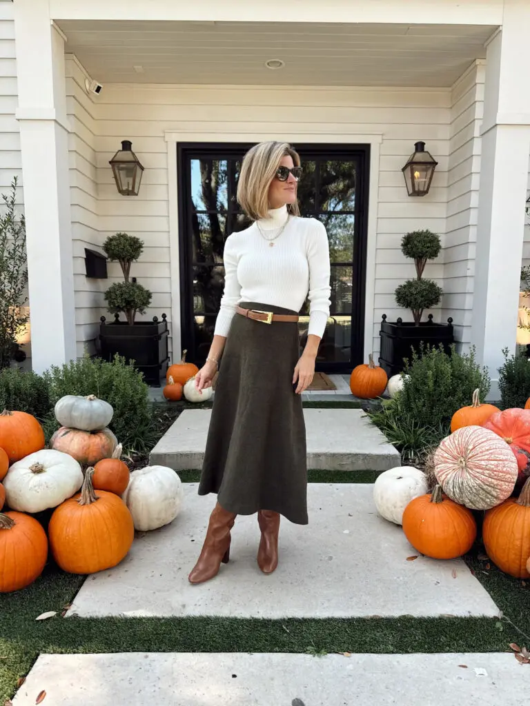 Woman in a white turtleneck, olive midi skirt and brown boots beside pumpkins