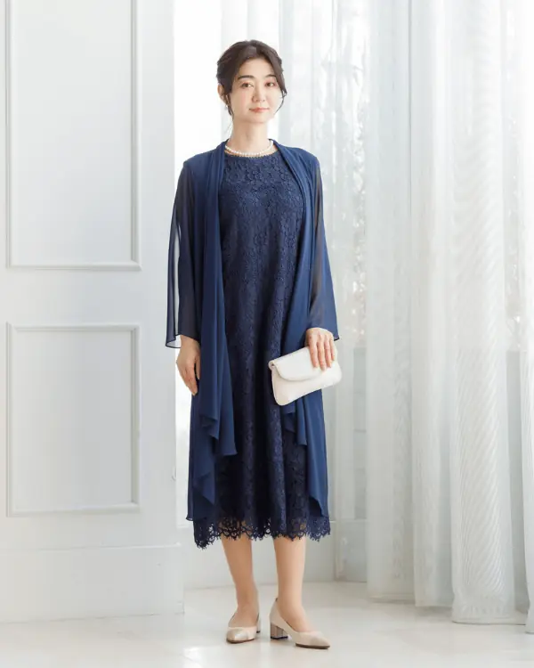 A mature woman wearing a navy lace midi dress with a matching chiffon shawl, pearl jewelry and a white clutch