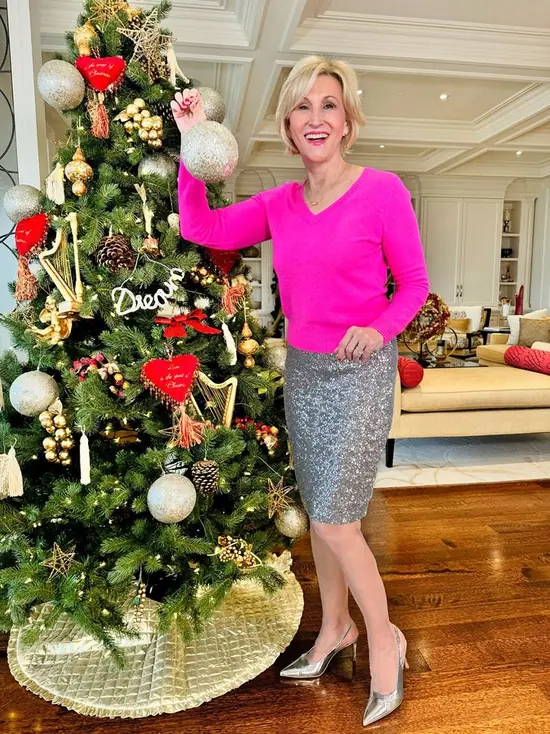 A woman over 50 wearing a bright pink cashmere sweater with a silver sequin pencil skirt beside a Christmas tree