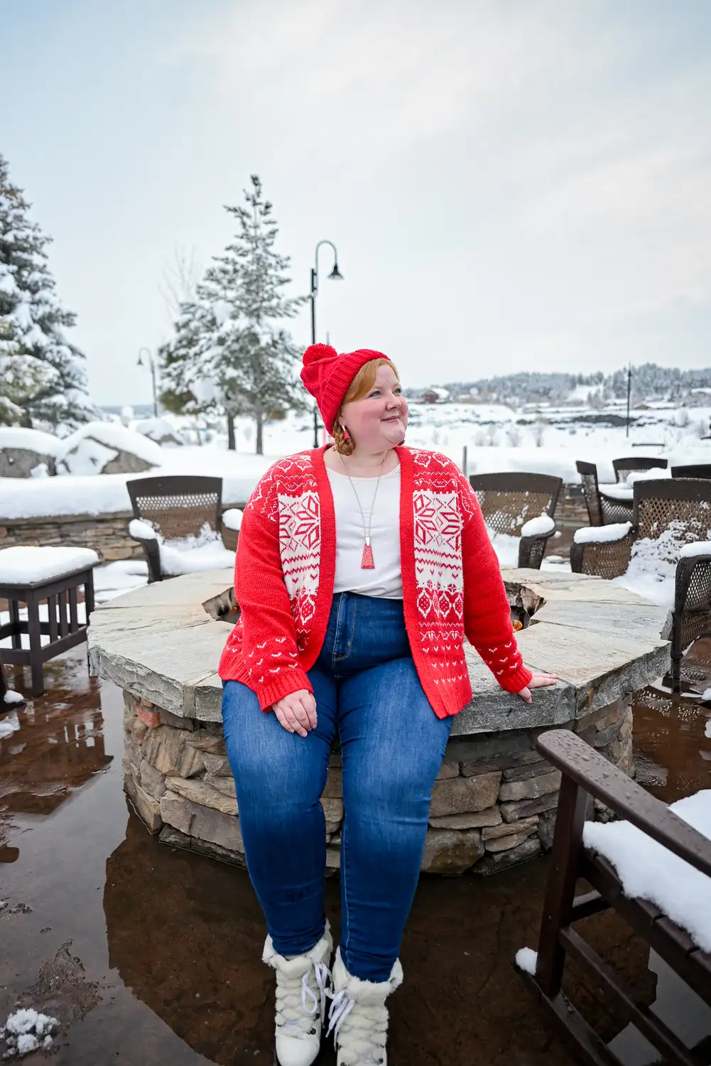Midsize woman wearing a red cardigan blue jeans knit hat and white snow boots outdoors