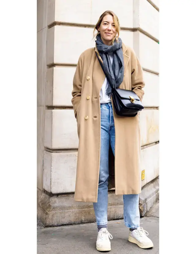 Woman over 50 wearing a camel coat with jeans, scarf, and white sneakers for winter travel