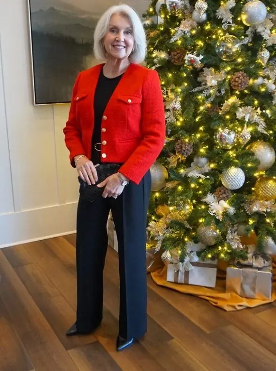Older woman wearing a red jacket with black wide leg trousers beside a Christmas tree