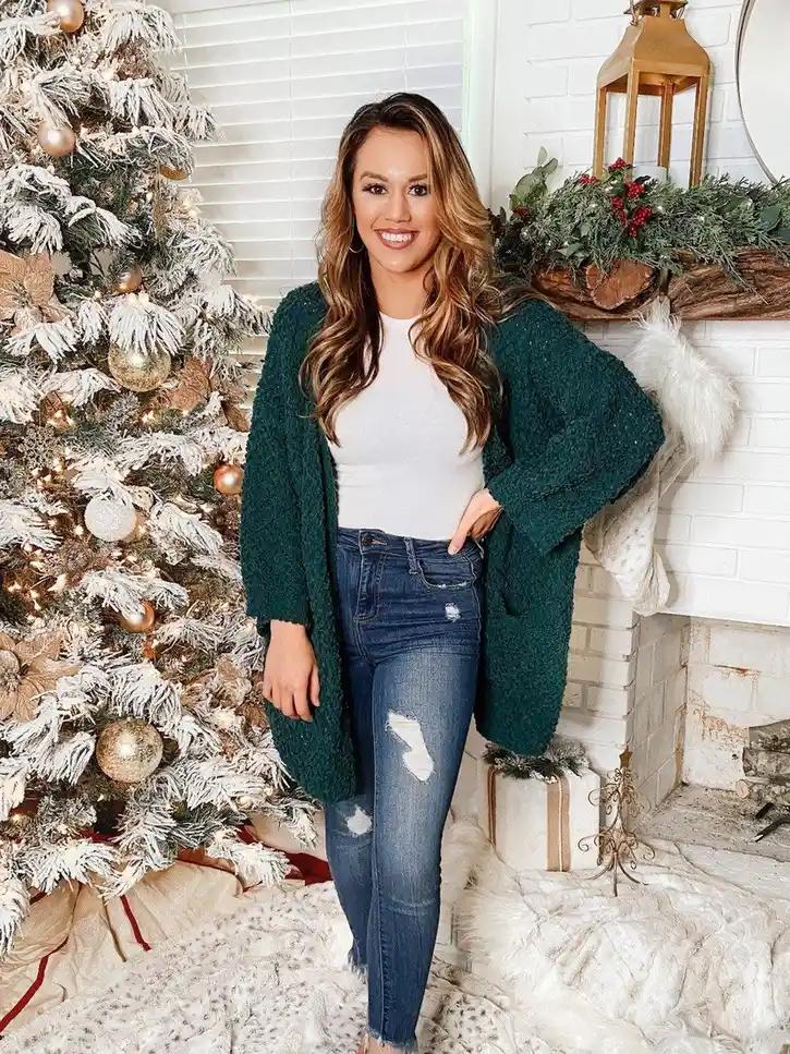 Woman wearing an emerald green cardigan with a white top and blue jeans beside a Christmas tree