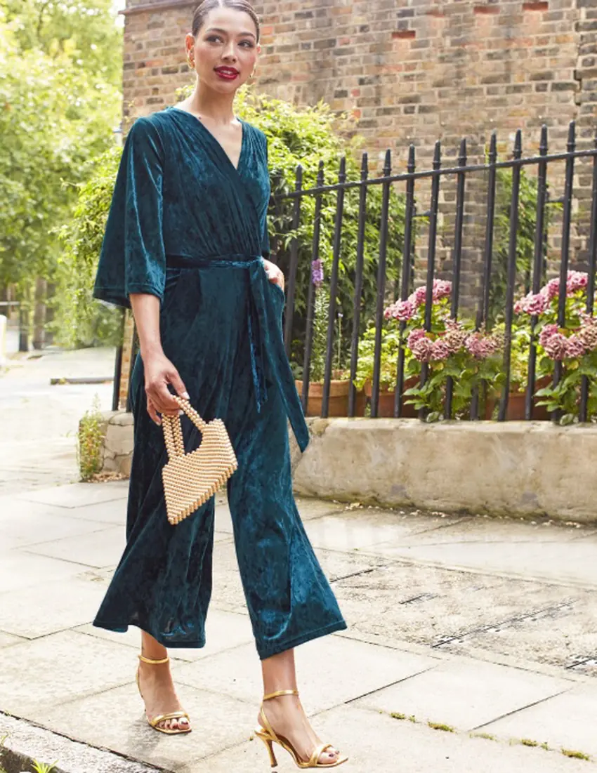 A woman wearing a teal velvet wide leg jumpsuit with a tie waist, gold heels and a beaded handbag