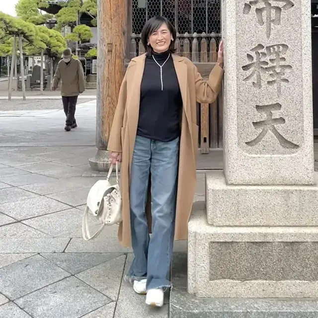 Woman in her 50s wearing a camel coat with a black knit, wide jeans, and white sneakers