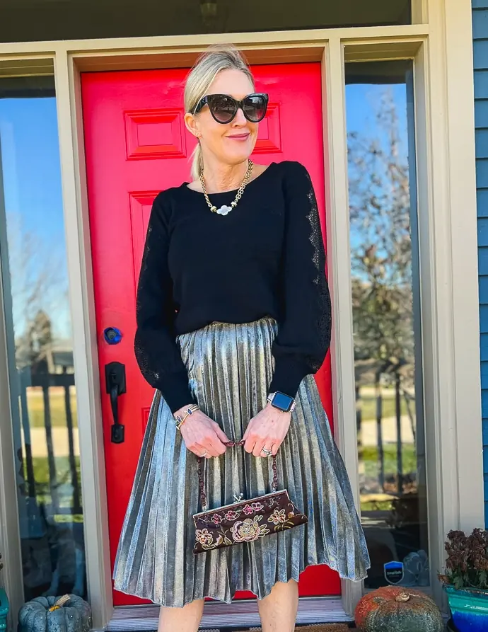 A silver haired woman wearing a black sweater with a silver metallic pleated midi skirt and festive accessories