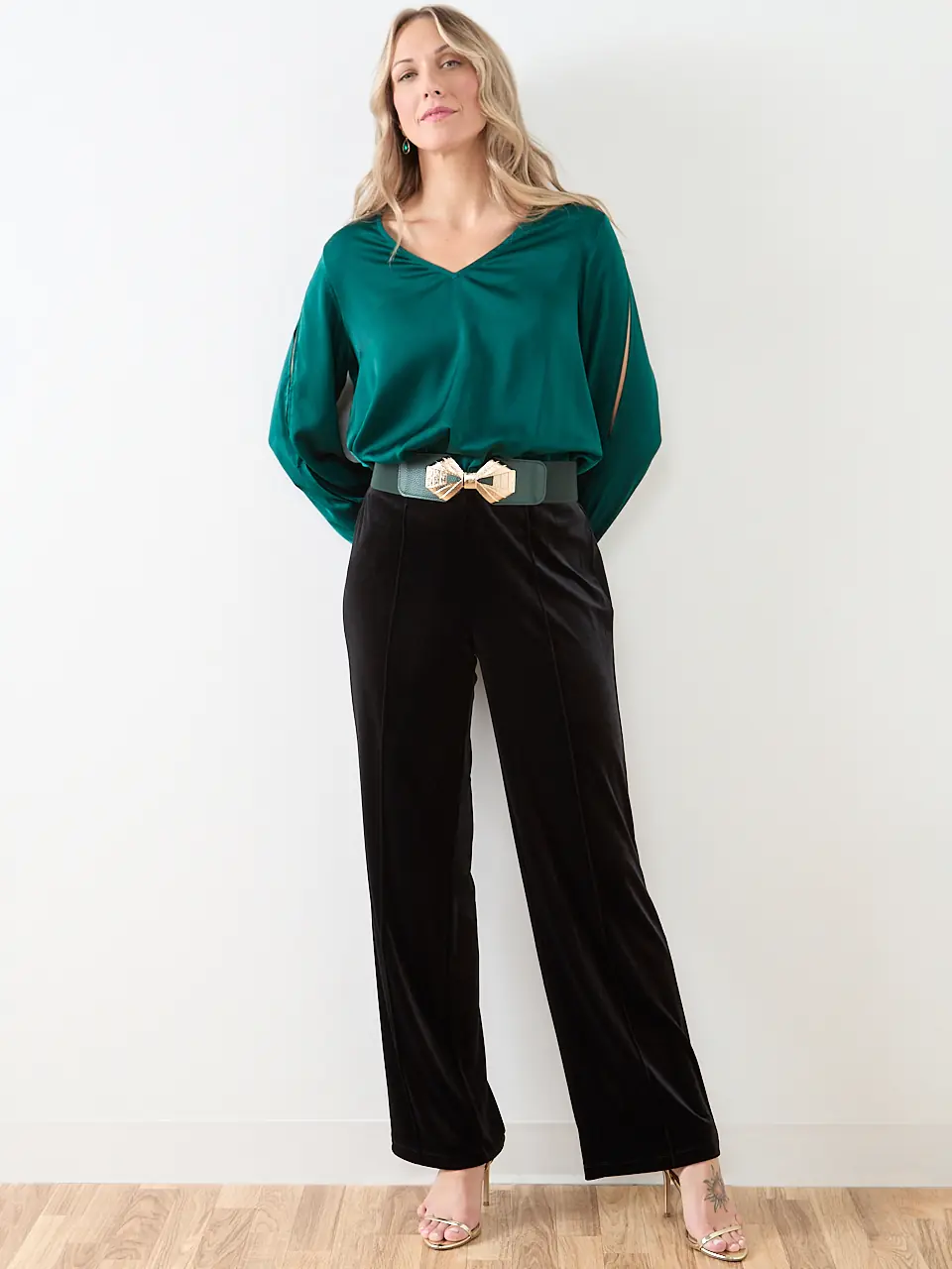 A woman wearing an emerald satin blouse tucked into black velvet wide leg trousers with a gold buckle belt