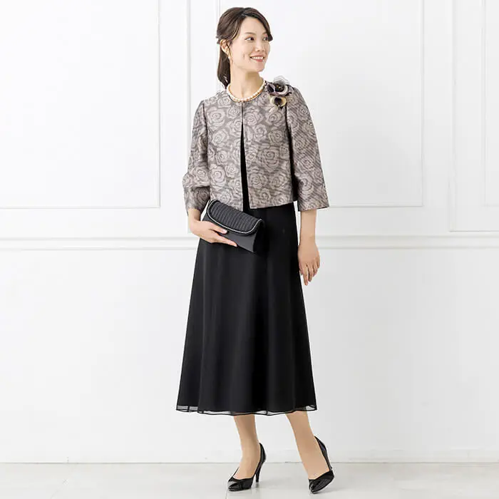 A mature woman wearing a bronze floral jacquard jacket over a black midi dress with pearl jewelry and black pumps