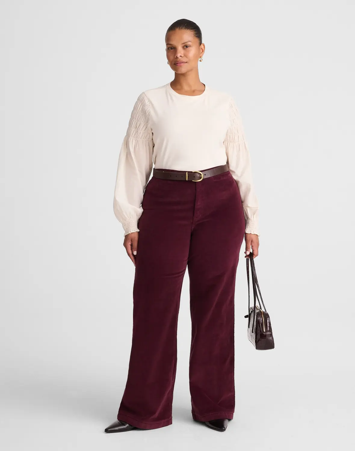A woman wearing a cream long sleeve blouse with burgundy wide leg corduroy pants and a slim belt