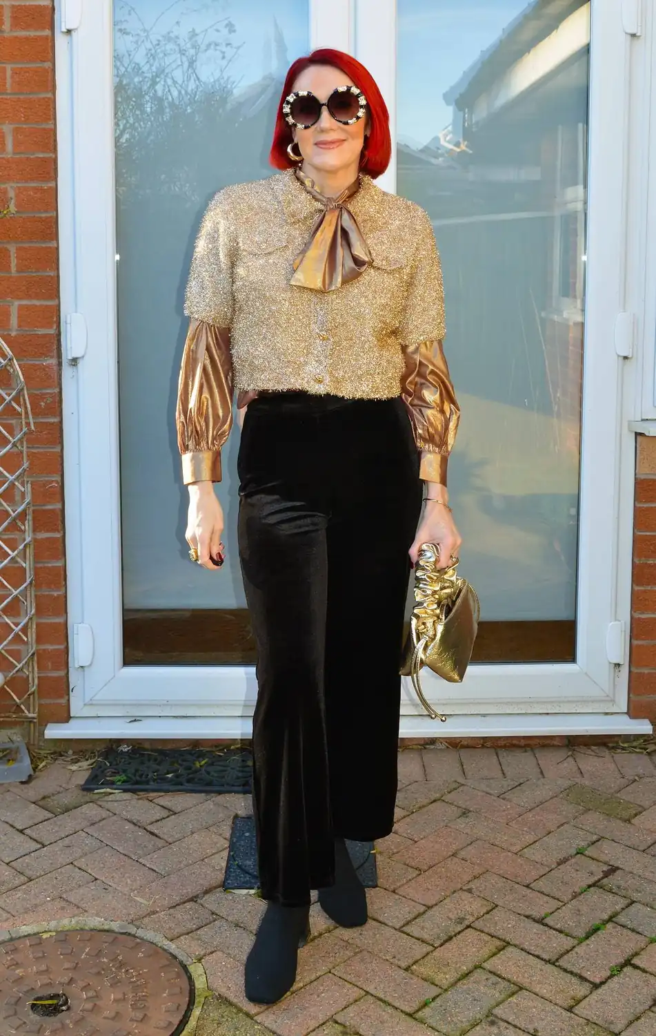 Red haired woman wearing a gold blouse and cardigan with black velvet trousers