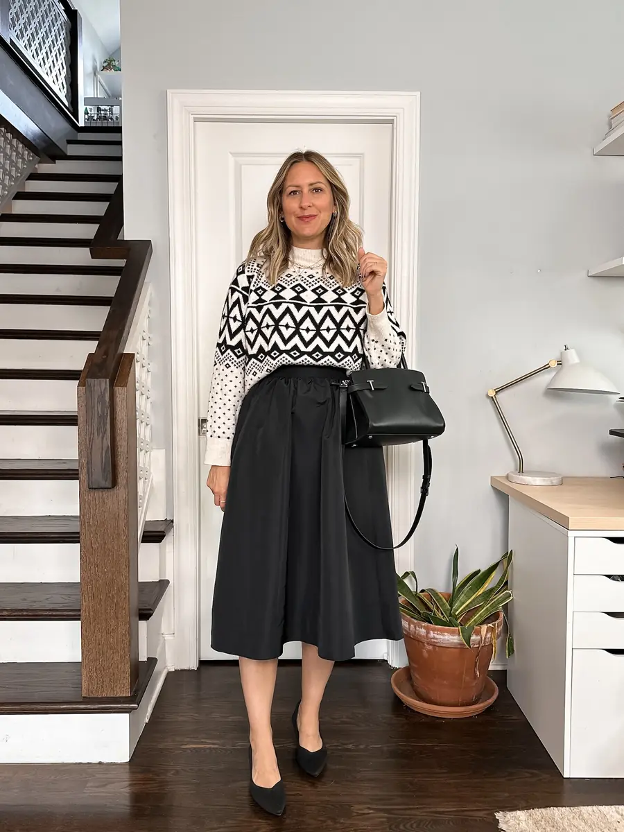 A woman wearing a black and white Fair Isle sweater with a flowing black midi skirt, pumps and a structured handbag