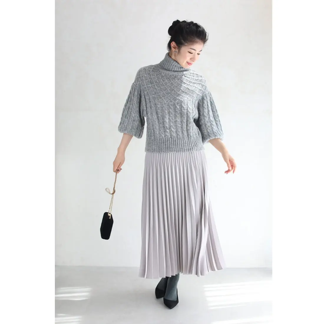 A woman wearing a soft grey cable knit sweater with a silver pleated midi skirt and black ankle boots