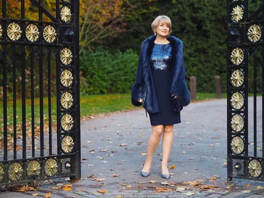 A stylish woman over 40 wearing a navy sequin dress with a matching fur trim coat at a formal winter venue