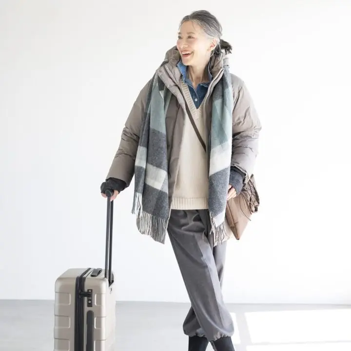 Woman over 60 wearing a taupe puffer, cream knit, grey trousers, scarf, and travel luggage