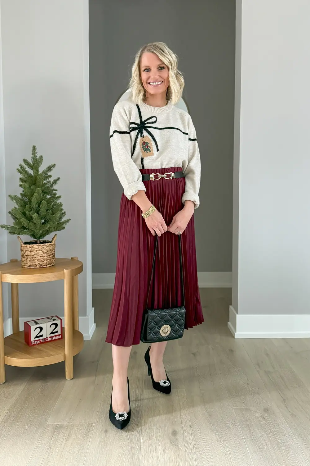 Woman wearing a cream holiday sweater with a burgundy pleated midi skirt and black pumps