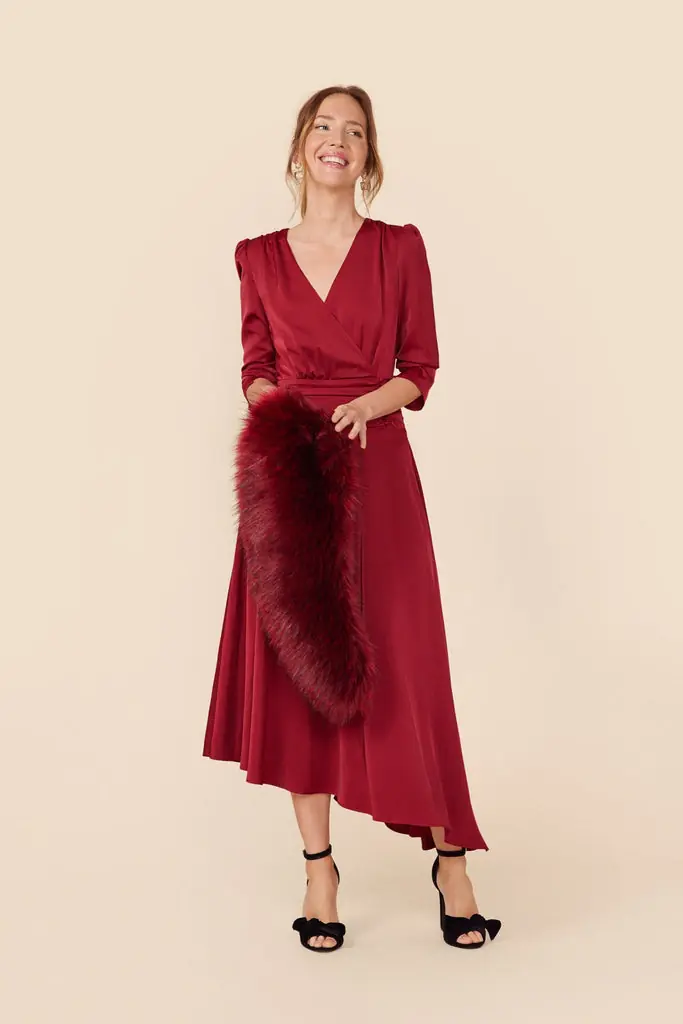 A woman wearing a burgundy wrap midi dress with an asymmetrical hem, black heels and a matching faux fur stole