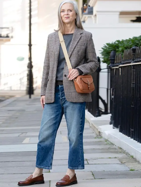 Mature woman wearing a plaid blazer with a grey sweater, jeans, loafers, and a crossbody bag