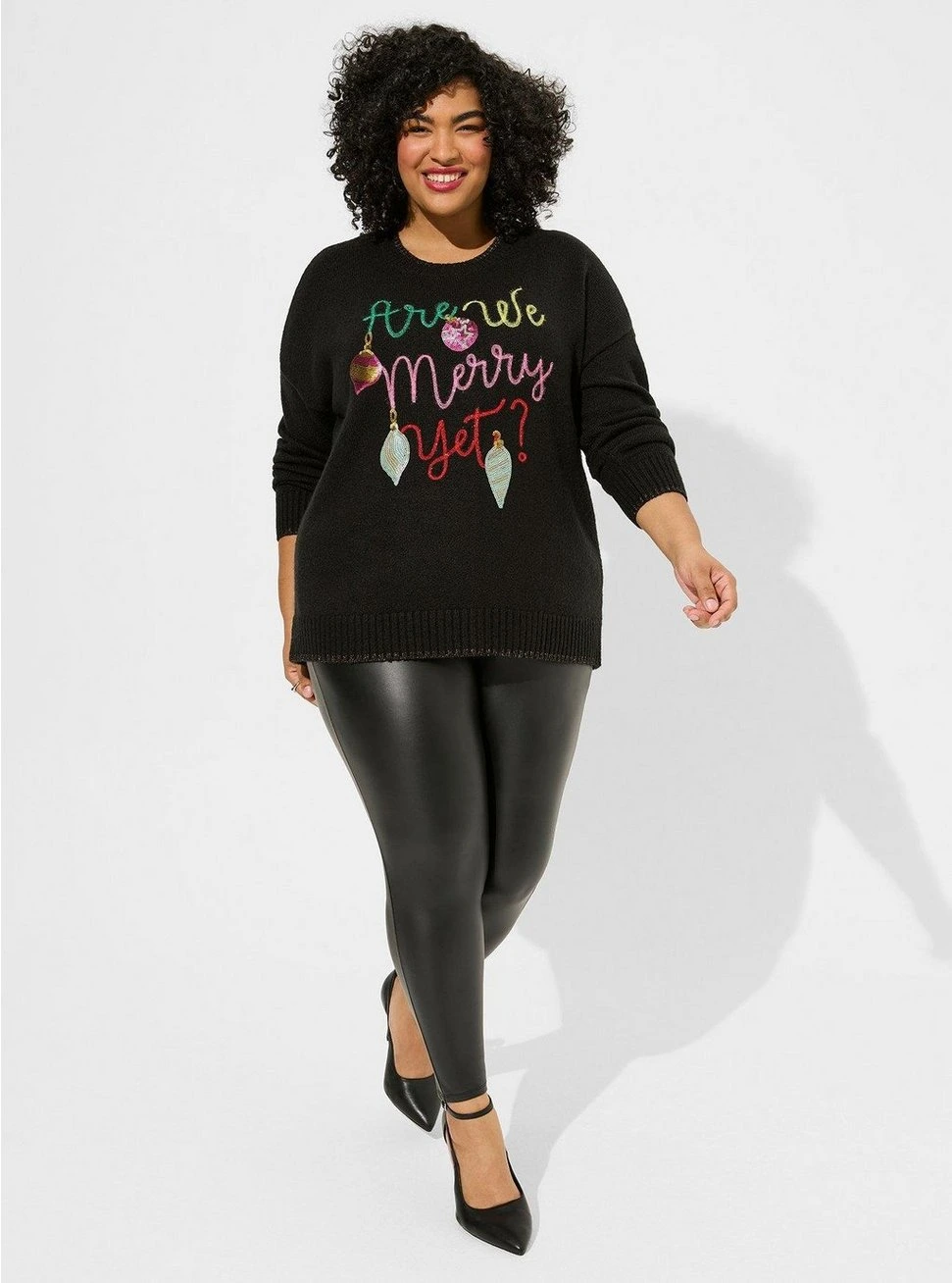 Curvy woman wearing a black holiday graphic sweater with faux leather leggings and pointed shoes