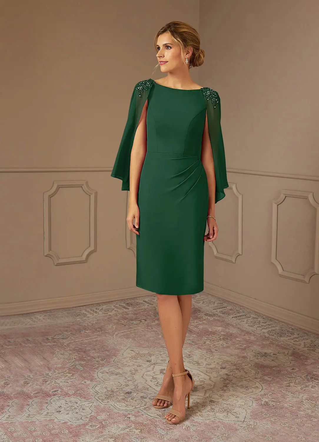 A woman wearing a dark green knee length wedding guest dress with flowing cape sleeves and nude heels