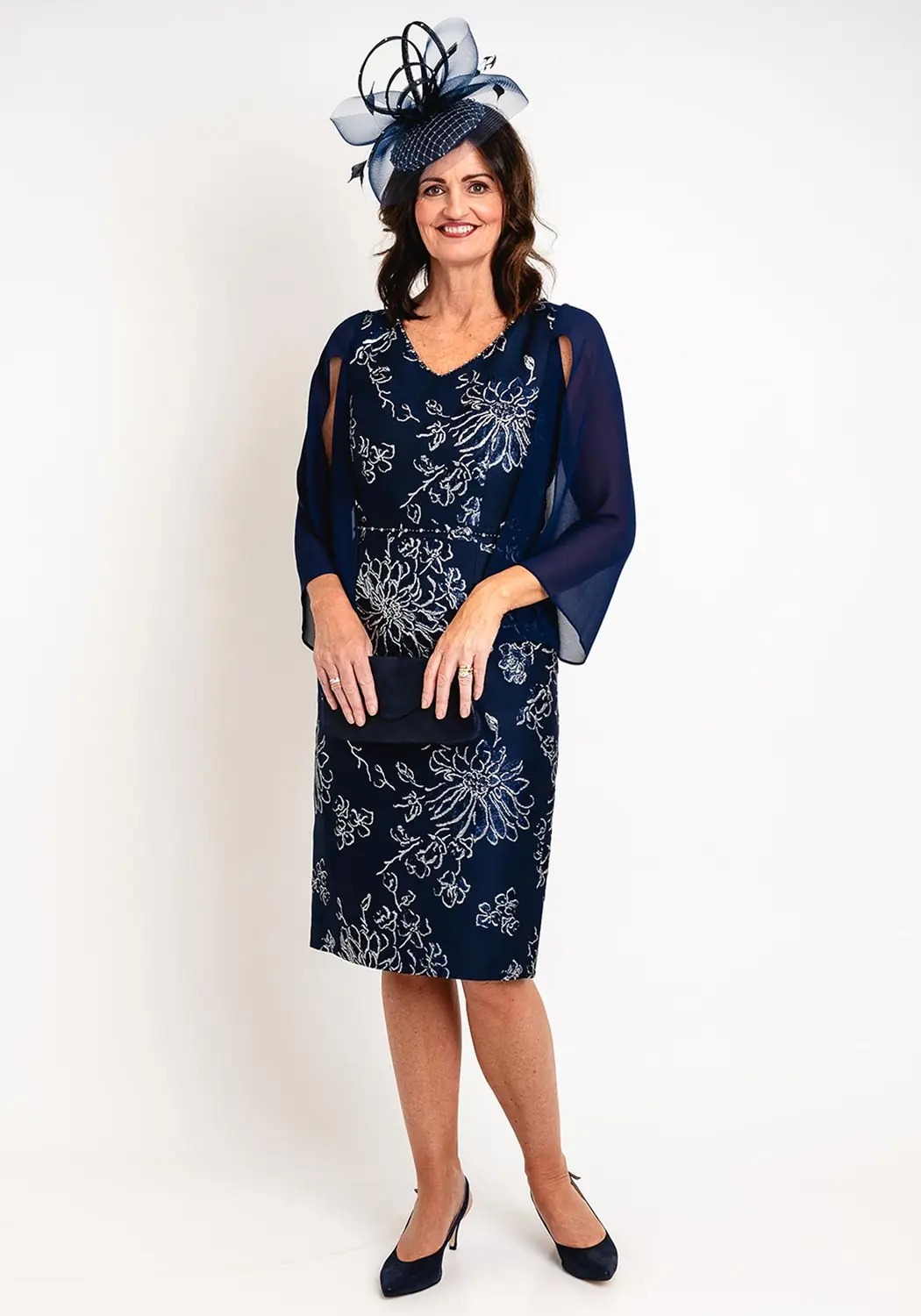 A mature woman wearing a navy floral jacquard midi dress with chiffon sleeves, a matching clutch and navy pumps