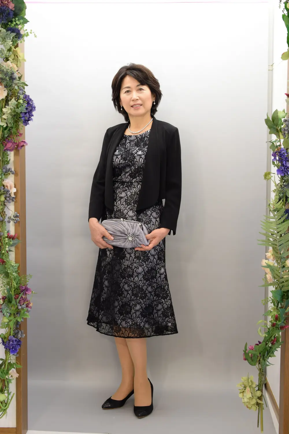 A woman over 50 wearing a black lace knee length dress with a tailored black velvet blazer, pearls and a grey clutch