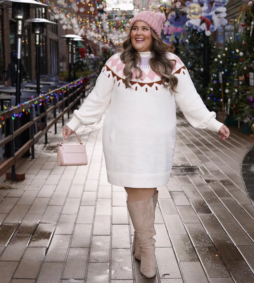 Midsize woman wearing a white Fair Isle sweater dress with a pink beanie and tall taupe boots