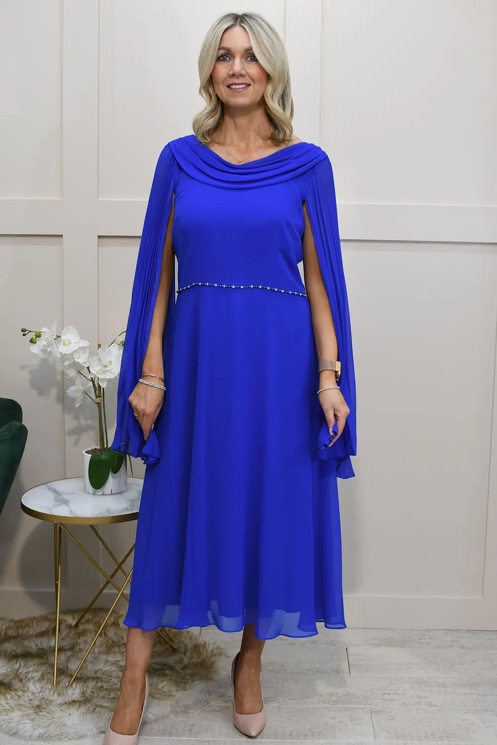 A mature woman wearing a cobalt blue midi dress with pleated cape sleeves, a slim jeweled belt and nude heels