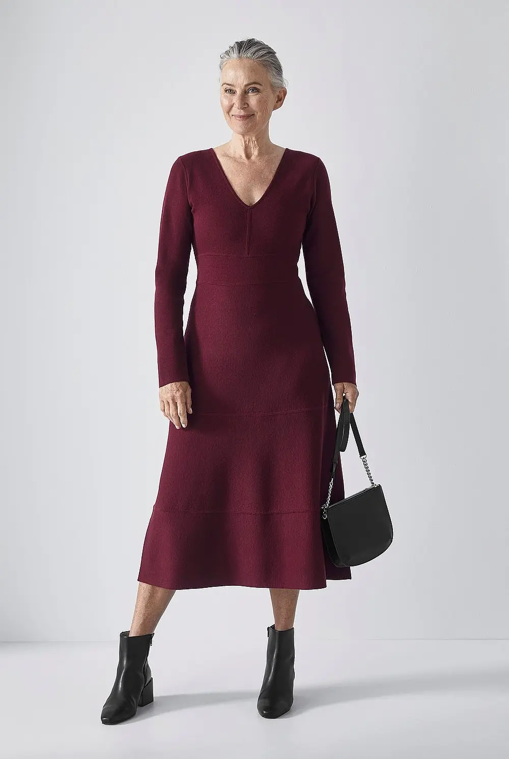 A silver haired woman wearing a cranberry long sleeve knit midi dress with black ankle boots and a black handbag